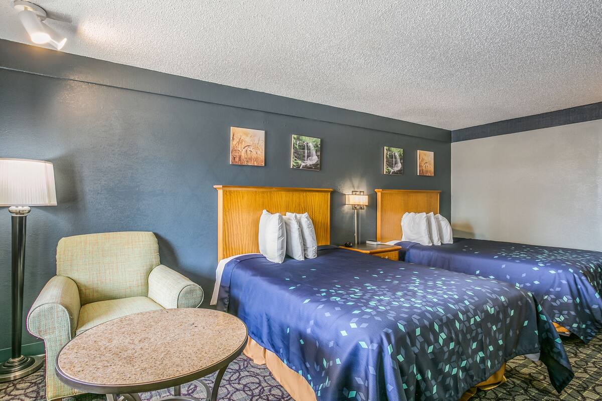 Habitación de hotel con dos camas, una silla, una mesita y obras de arte en la pared.