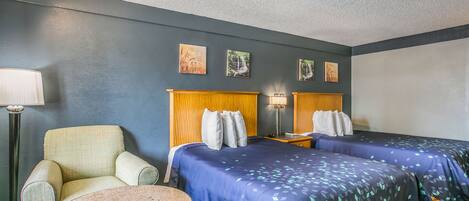 A hotel room with two beds, a chair, a small table, and artwork on the wall.