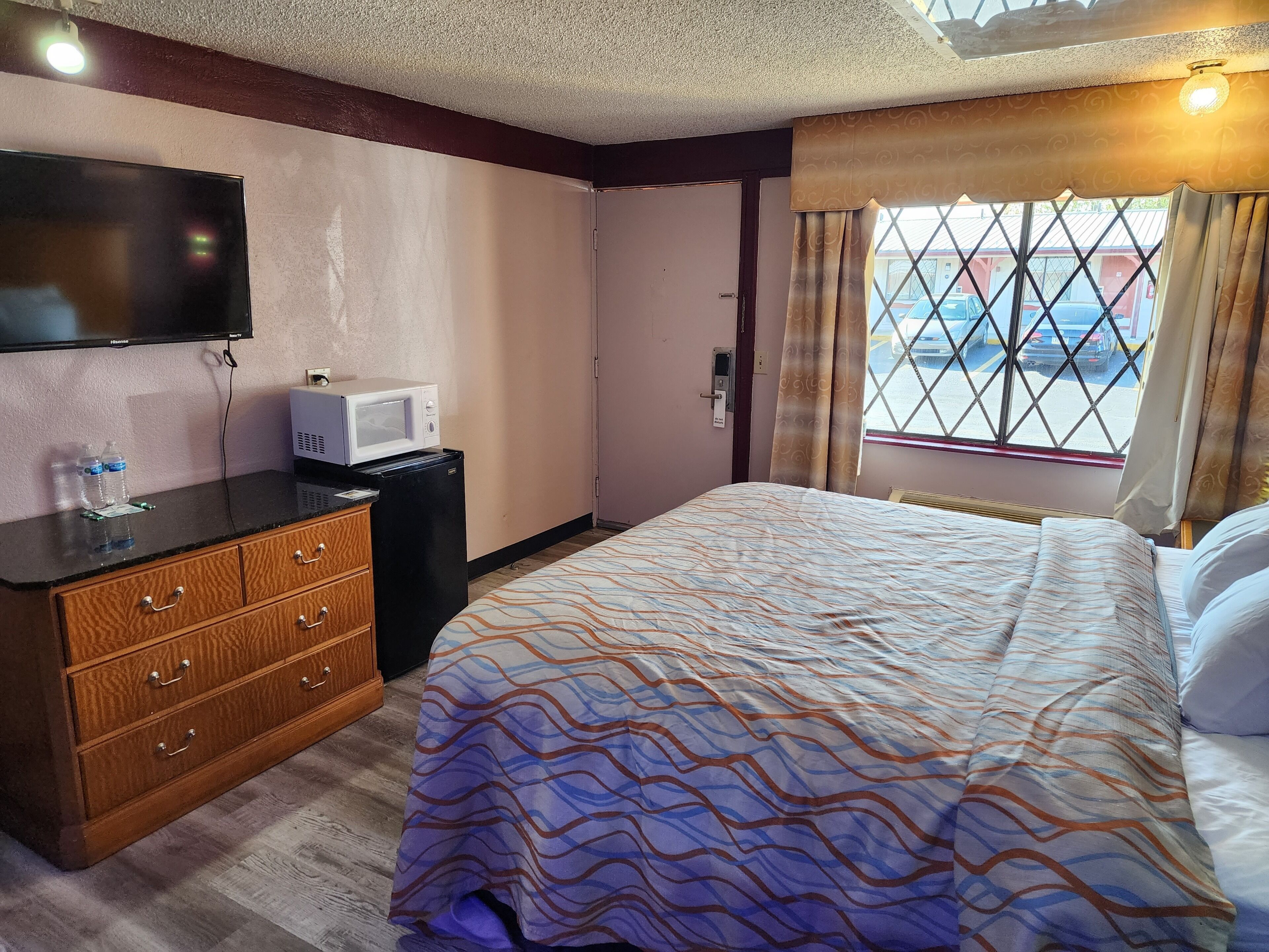 Habitación de hotel con cama, televisor, microondas, refrigerador pequeño, cómoda de madera y ventana con vista a coches estacionados.