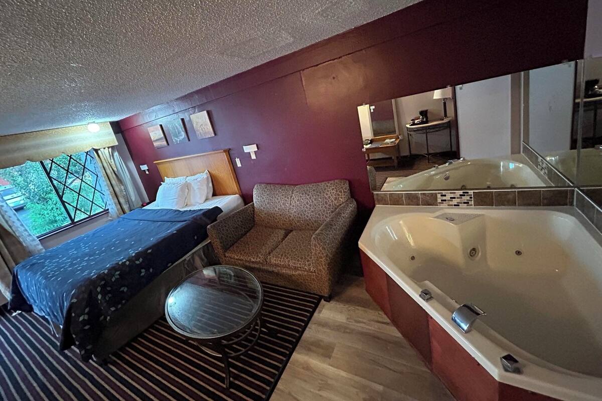 Habitación de hotel con cama, sofá, una mesita redonda, una bañera grande y ventana con vista a follaje.