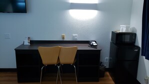 A small kitchenette with a microwave, refrigerator, and a flat-screen TV mounted on the wall. There are two chairs in front of a desk with a telephone and a small table with a box. - Motel 6 Sedalia, MO (Sedalia)