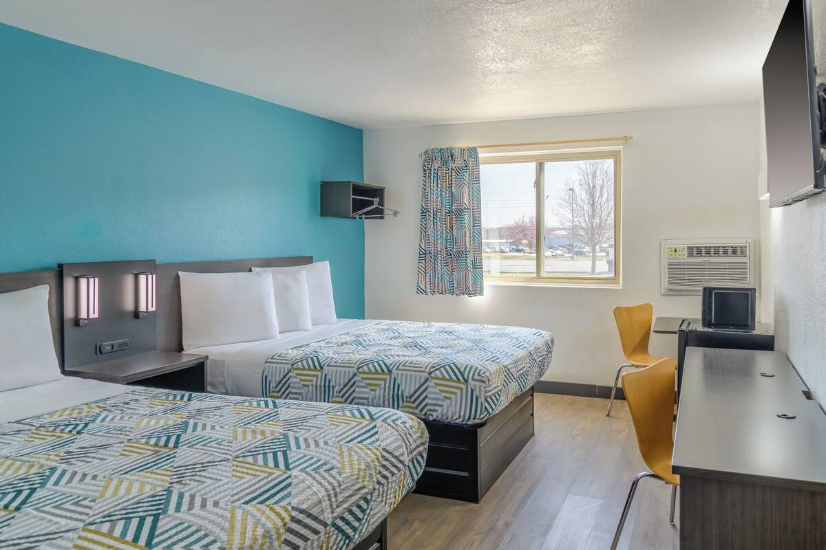 A hotel room with two beds, a desk, a chair, a TV, and a window with a view of the outside.