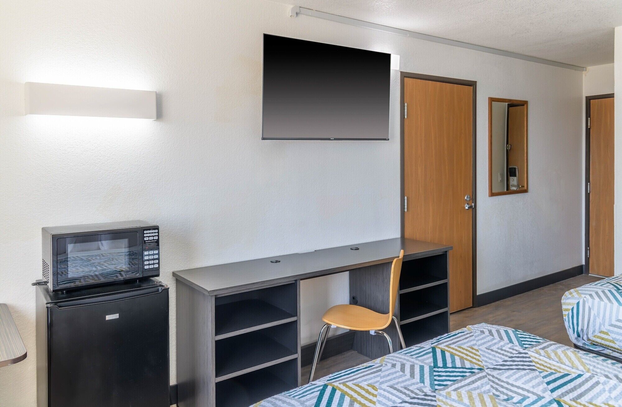 Une chambre d’hôtel compacte comprenant un lit, un bureau avec une chaise, un petit réfrigérateur, un téléviseur à écran plat fixé au mur et une porte dotée d’un miroir.