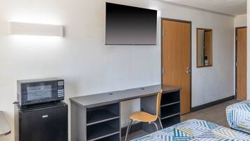 A compact hotel room with a bed, a desk with a chair, a small refrigerator, a flat-screen TV mounted on the wall, and a door with a mirror.