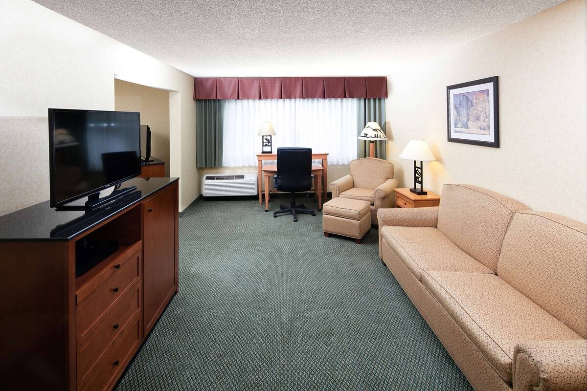 A hotel room with a flat-screen TV, a wooden cabinet, a beige sofa, a small desk with a chair, and a framed picture on the wall.