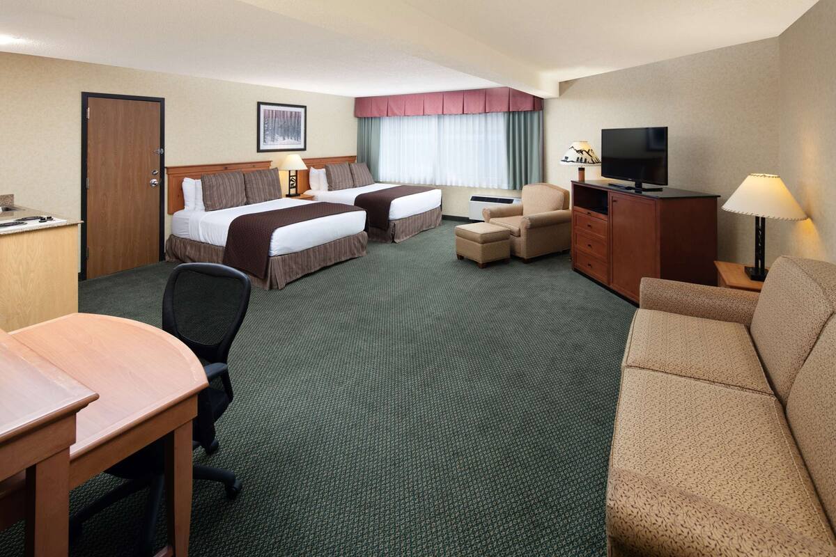 A hotel room with two beds, a desk, a chair, a sofa, a TV, and a lamp.