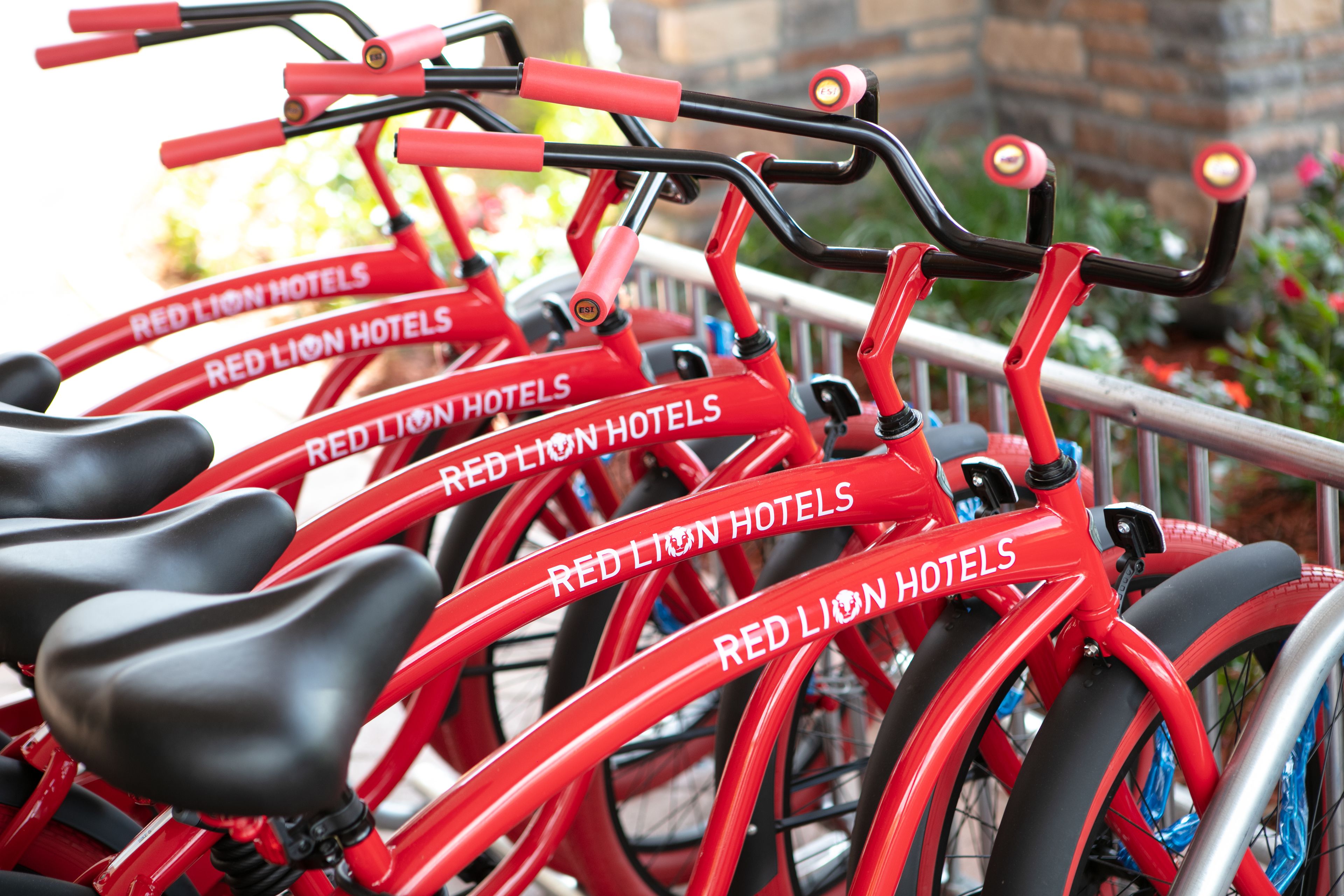 Une rangée de vélos rouges portant la marque « RED LION HOTELS », stationnés dans un espace extérieur agrémenté d’une rambarde en métal et d’une végétation en arrière-plan.
