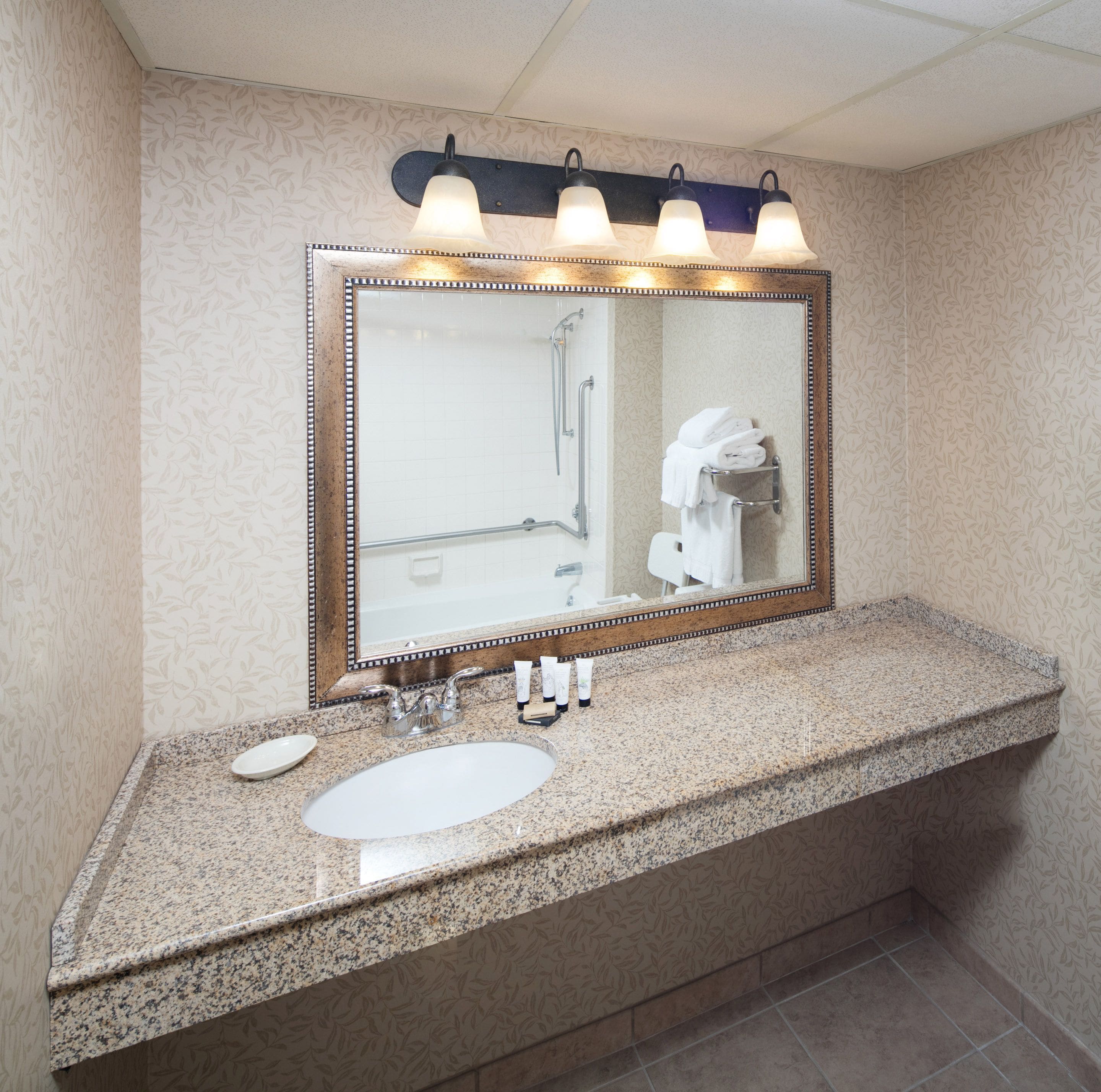 A bathroom with a granite countertop, a white sink, a large mirror with lights, and a towel rack.