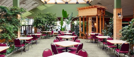 Un comedor interior con mesas y sillas, un techo alto con tragaluces y plantas verdes.