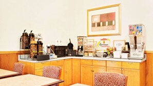 A breakfast bar with coffee machines, a toaster, and a refrigerator. There are wooden cabinets and patterned chairs.