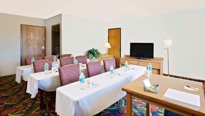 A hotel conference room with a long table, several chairs, a television, and a desk with a lamp and a basket of pens.