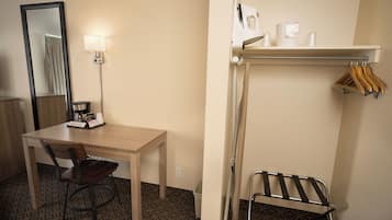 Een hotelkamer met een houten bureau, een stoel, een spiegel, een lamp, een plank met kledinghangers en een opklapbare stoel.