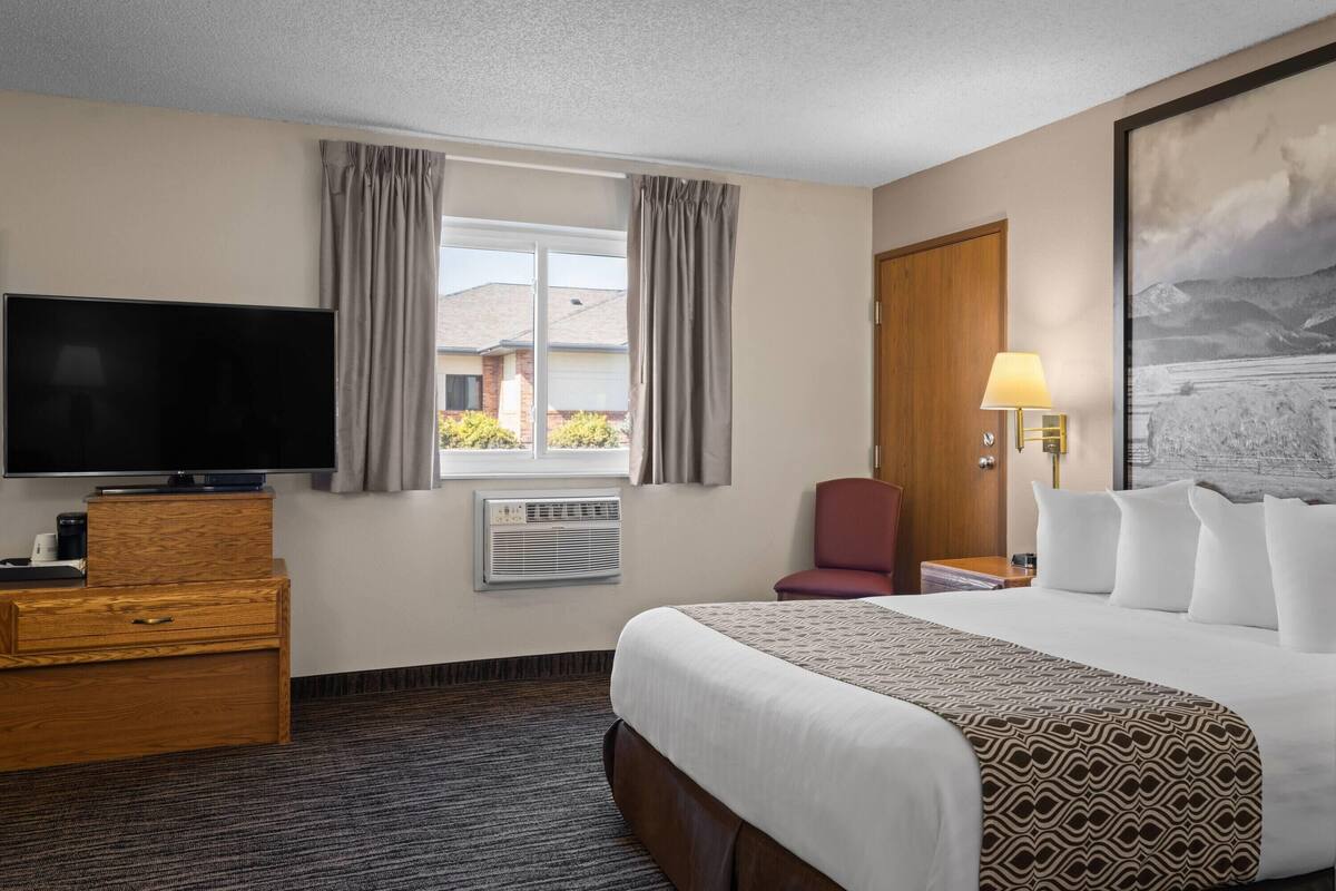 Een hotelkamer met een groot bed, een televisie op een houten dressoir, een stoel, een bureau met een lamp en een raam met gordijnen.