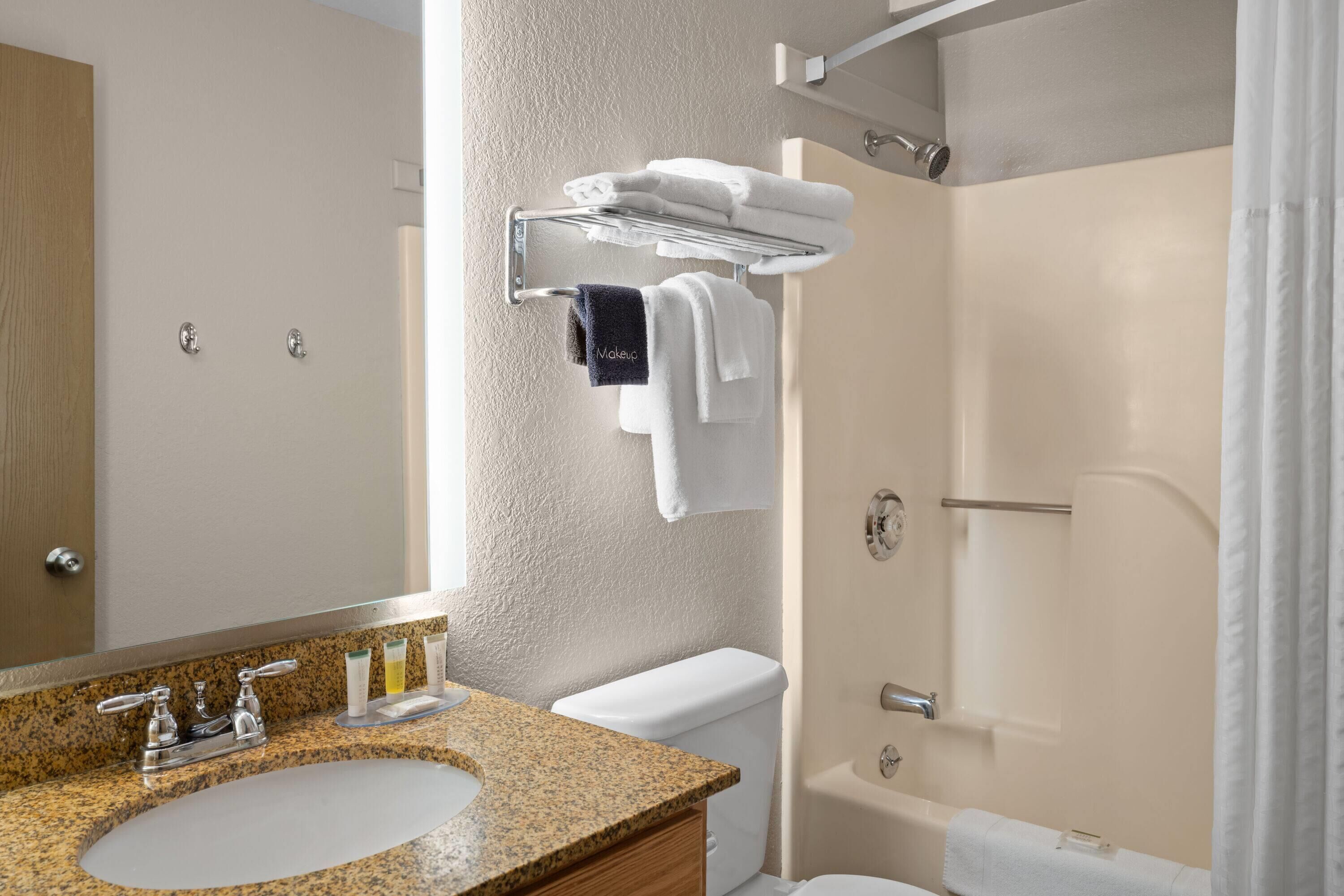 A bathroom with a granite countertop sink, a mirror, a towel rack with white and dark blue towels, a toilet, and a shower area.