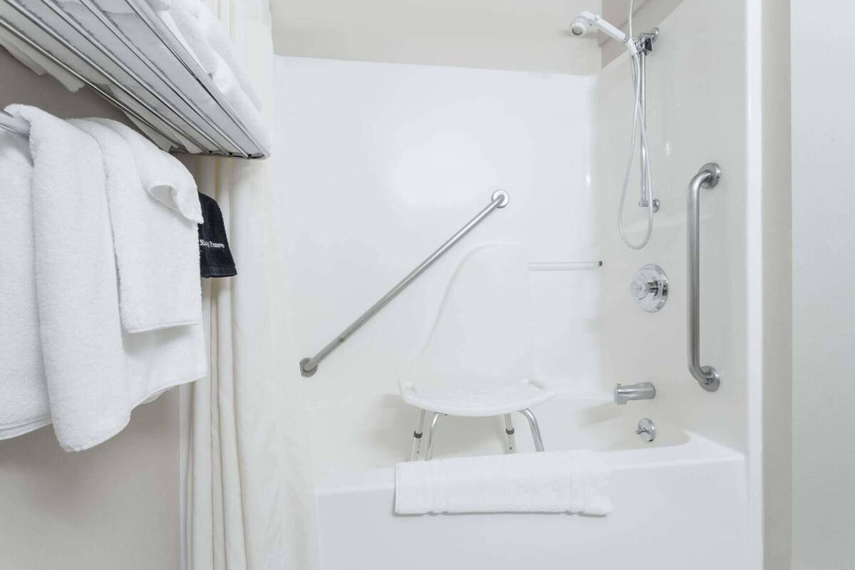 Een badkamer met een wit bad, een douche met een handdouche en een handgreep. Er hangen witte handdoeken aan een rek.