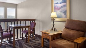 Una habitación de hotel con una mesa de comedor de madera, cuatro sillas, un sillón de cuero marrón, una pequeña mesita de noche con una lámpara y un cuadro con un paisaje en la pared.