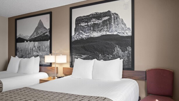 Habitación de hotel con dos camas, una silla roja, una lámpara y dos cuadros con paisajes de montañas.