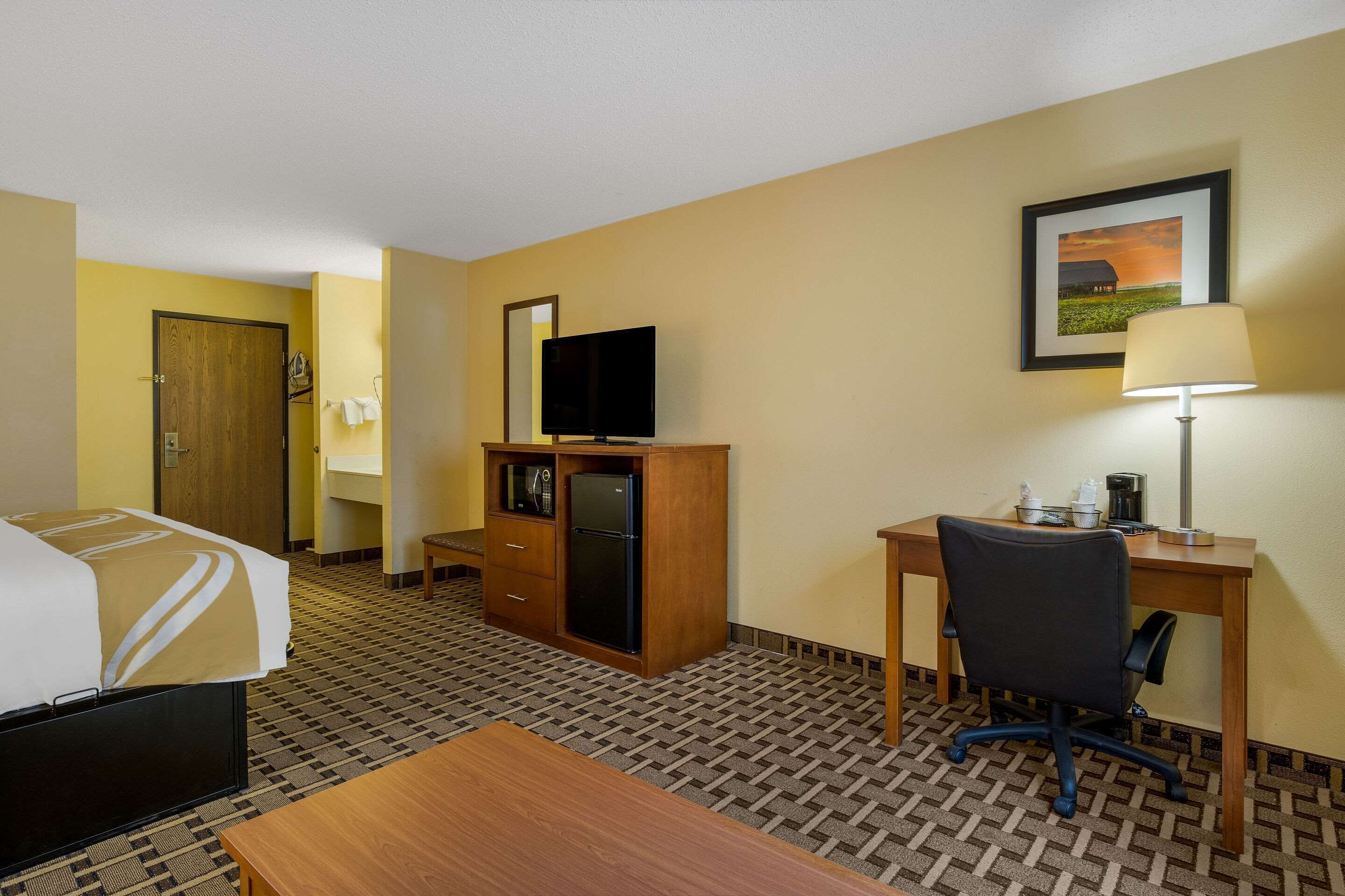 Habitación de hotel con cama, escritorio, silla, televisor, refrigerador, lámpara y un cuadro en la pared.