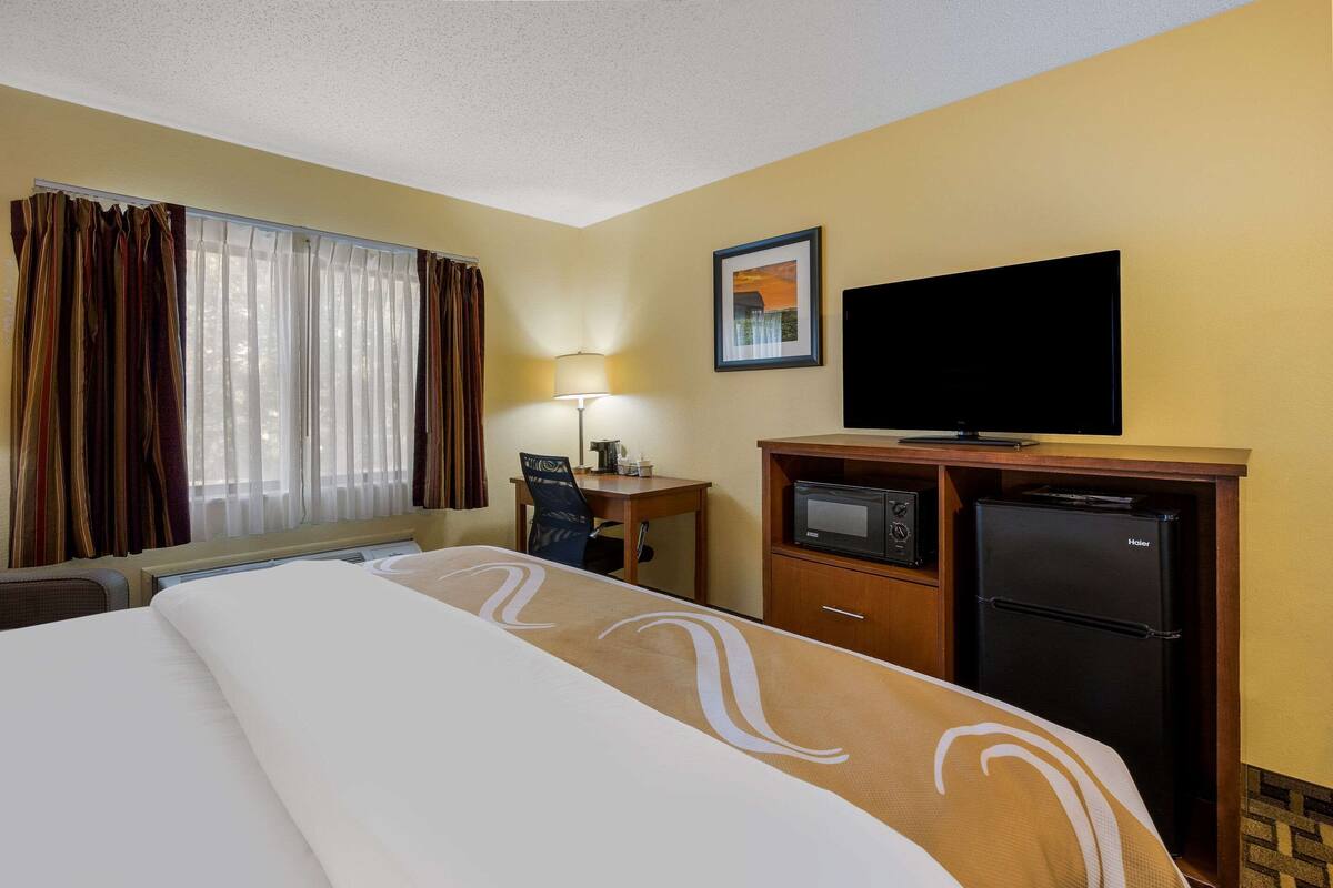 A hotel room with a bed, a desk, a television, a refrigerator, and curtains.