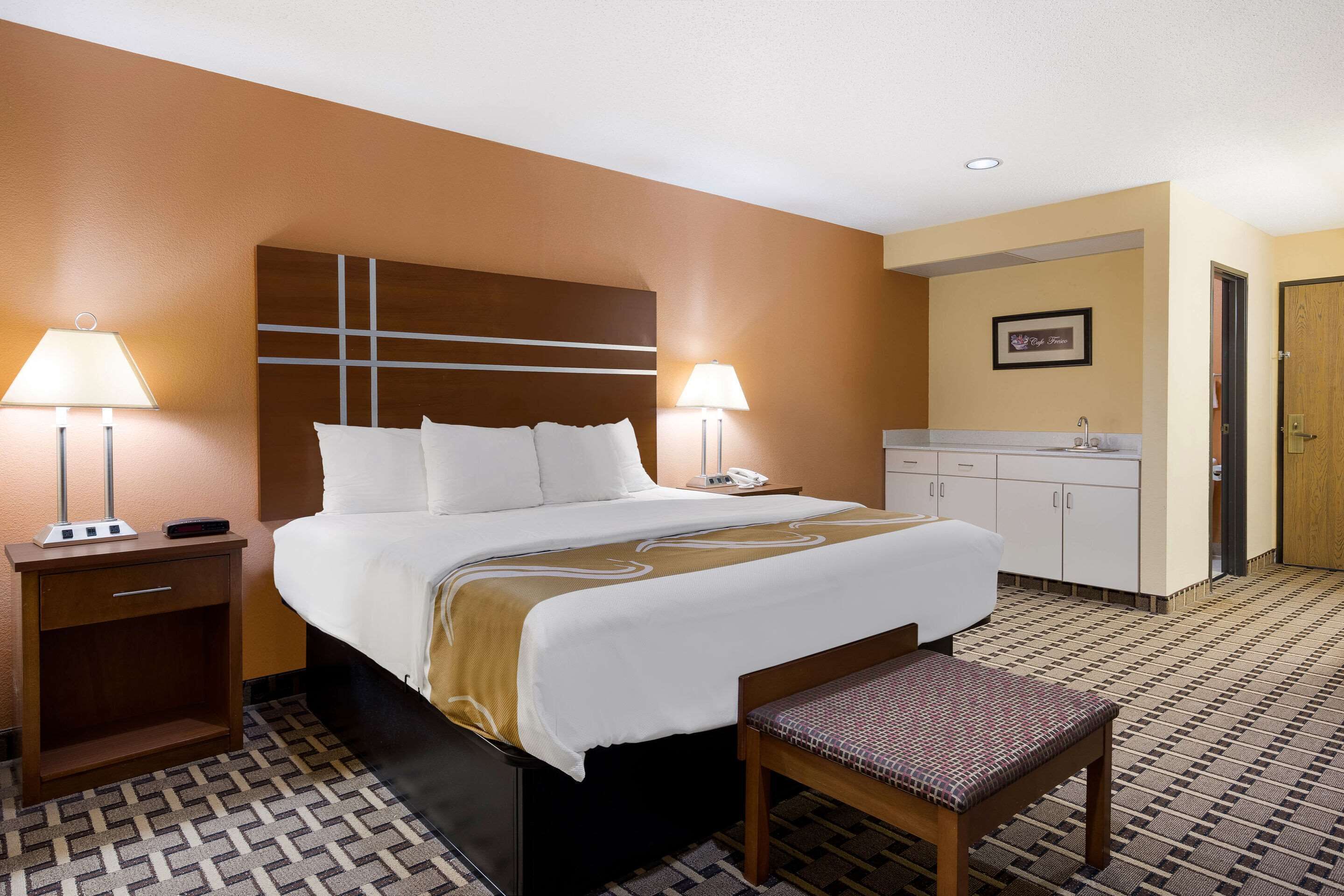 Una habitación de hotel con una cama grande, lámparas de noche, una mesita, un banco, una kitchenette y una puerta.