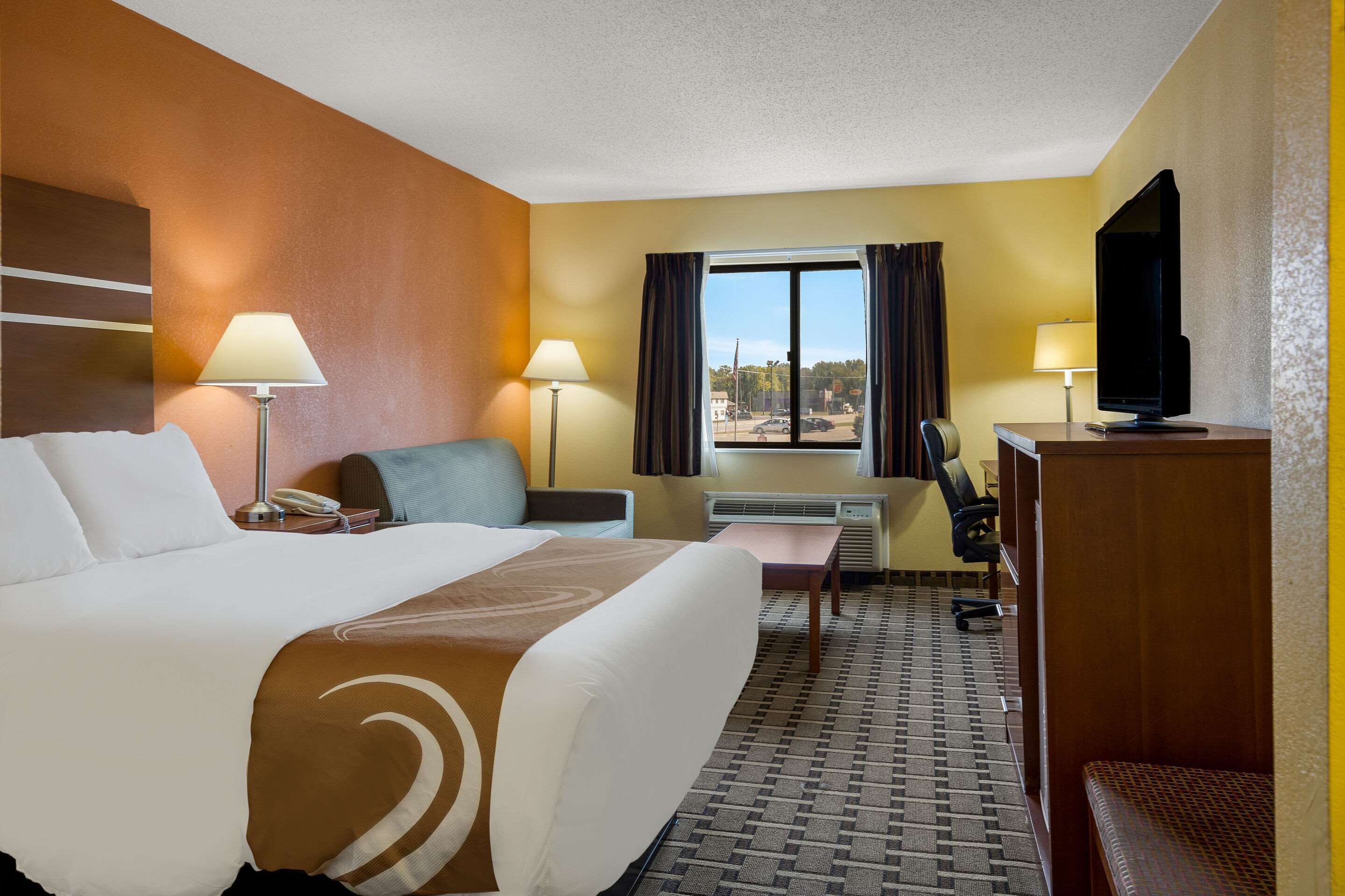 Une chambre d’hôtel avec un grand lit, deux tables de chevet avec des lampes, un bureau avec une chaise, une télévision et une fenêtre avec des rideaux.