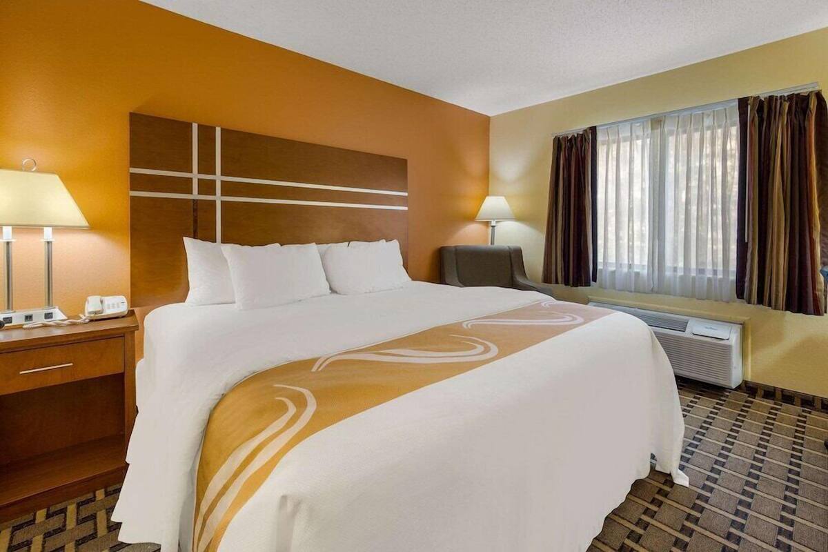A hotel room with a large bed, two bedside lamps, a desk with a telephone, and an air conditioning unit.