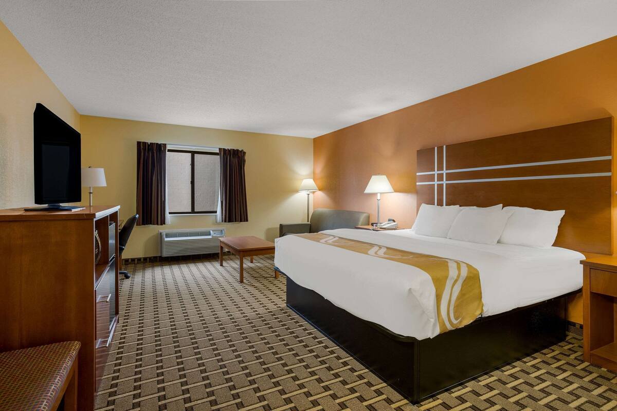 A hotel room with a large bed, a desk with a television, a chair, a lamp, and a window with curtains.