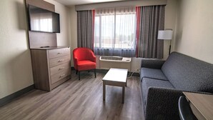 A hotel room with a flat-screen TV mounted on the wall, a red armchair, a grey sofa, a small white table, and a standing lamp. - Best Western Helena/Capitol Hotel (Helena)