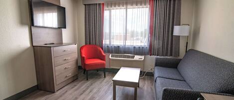 A hotel room with a flat-screen TV mounted on the wall, a red armchair, a grey sofa, a small white table, and a standing lamp.