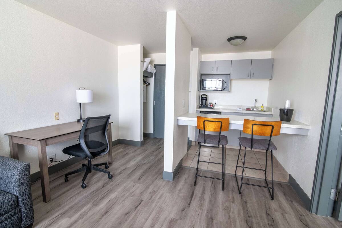 A compact living space with a kitchenette, a dining area with two bar stools, a desk with a chair, and a lamp.