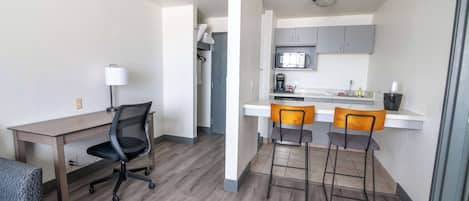 A compact living space with a kitchenette, a dining area with two bar stools, a desk with a chair, and a lamp.