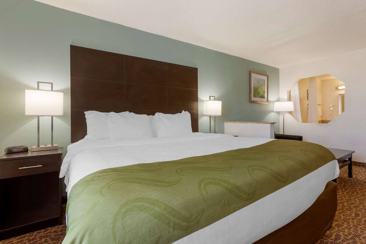 A hotel room with a large bed, two bedside tables with lamps, a nightstand, a mirror, and a carpeted floor.