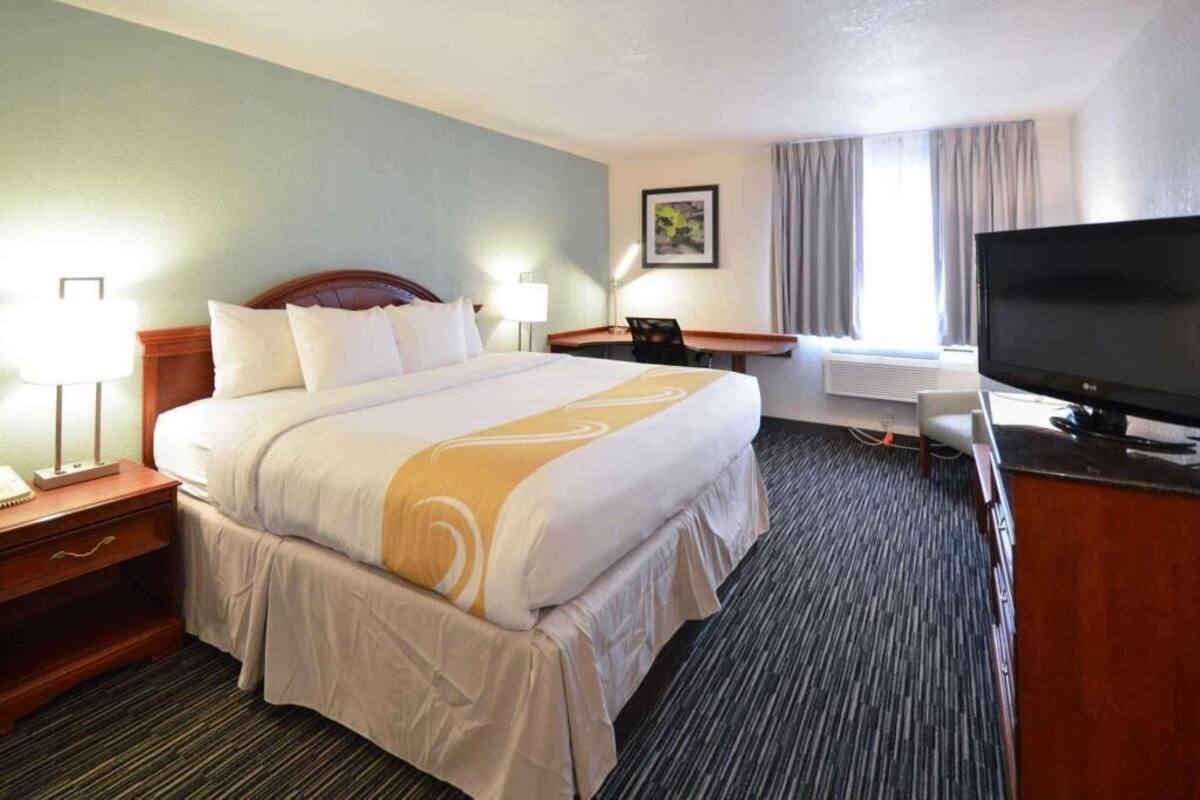 A hotel room with a large bed, a desk with a flat-screen TV, a chair, a lamp, and a window with curtains.
