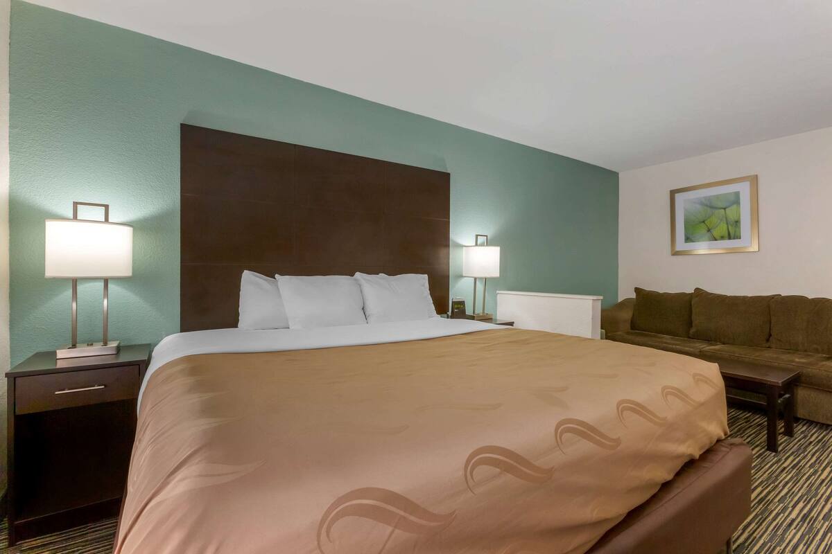 A hotel room with a large bed, two bedside tables with lamps, a brown sofa, and a framed picture on the wall.