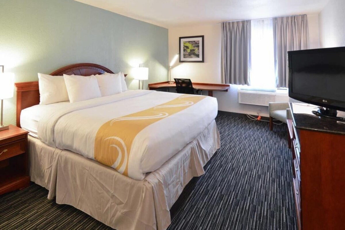 Quarto de hotel com cama grande, escrivaninha com cadeira, televisão e janela com cortinas.