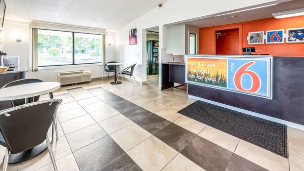 A motel lobby with a reception desk, a small seating area, and a view of the outdoors through large windows.