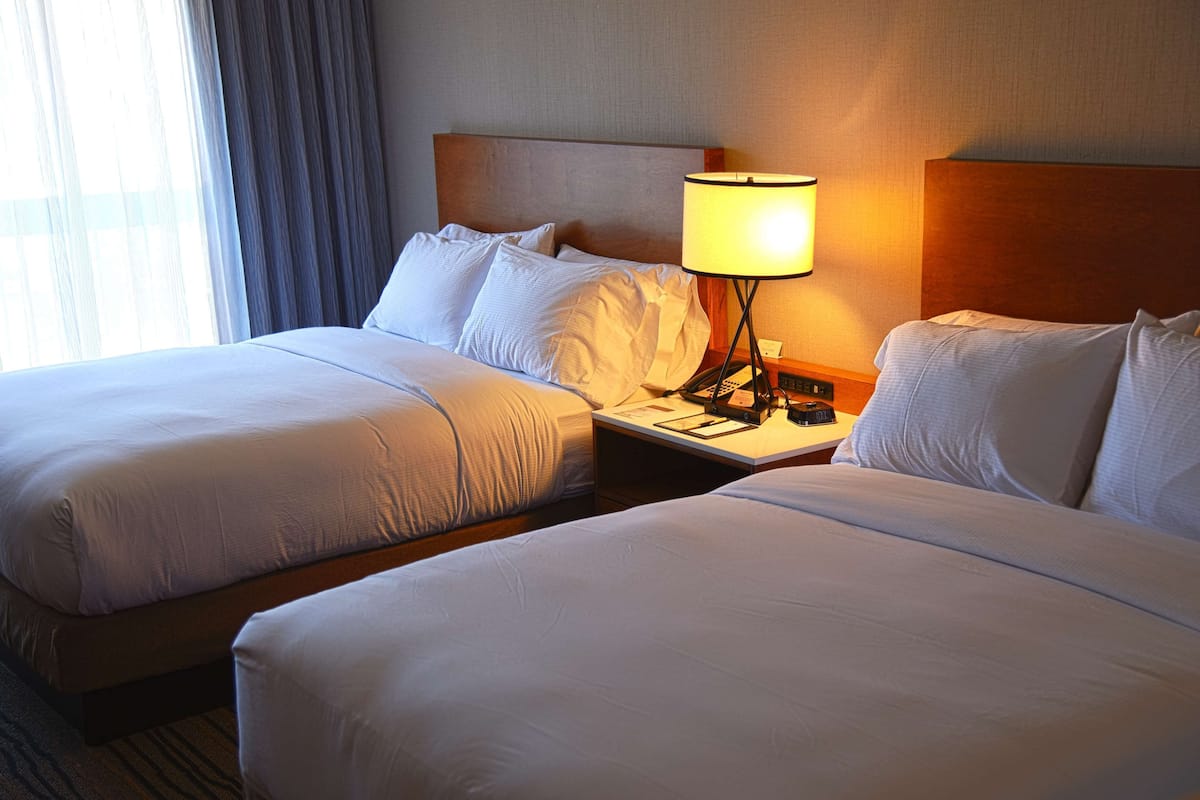 A hotel room with two beds, a nightstand with a lamp, and a window with curtains.