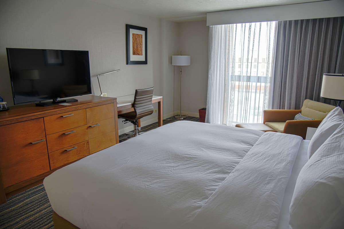 A hotel room with a large bed, a desk with a flat-screen TV, a chair, a lamp, and a window with curtains.