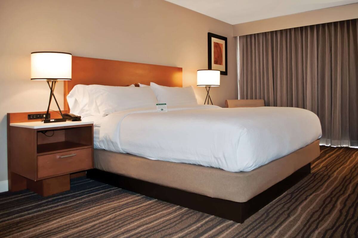 A hotel room with a large bed, two bedside tables with lamps, a desk with a chair, a framed picture on the wall, and a window with curtains.