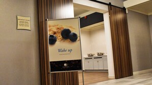 Un couloir d’hôtel avec un panneau indiquant le lieu du petit-déjeuner, une affiche faisant la publicité d’un buffet, et une cuisine visible au fond.