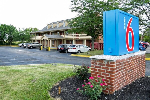 A motel with a large blue sign displaying the number 6. There are several cars parked in front, and the building has a traditional architectural style with a sloped roof and multiple windows. - Motel 6 - Dayton, OH (Dayton)