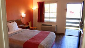 Habitación de hotel con cama, una silla, escritorio, lámpara, ventana con cortinas y puerta corrediza que da a un balcón.