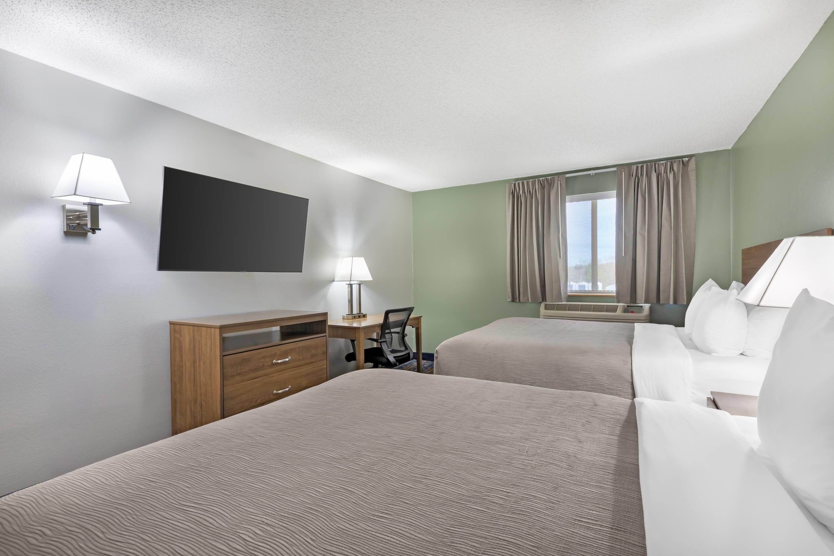 Ein Hotelzimmer mit zwei Betten, einem Schreibtisch, einem Sessel, einem Flachbildfernseher und einem Fenster mit Vorhängen.