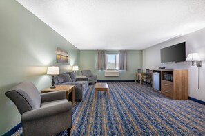 A hotel room with a sofa, armchair, coffee table, flat-screen TV, mini-fridge, microwave, and a large window with curtains.
