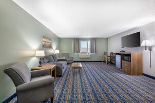 Habitación de hotel con sofá, sillón, mesa de centro, televisor de pantalla plana, mini refrigerador, microondas y un gran ventanal con cortinas.