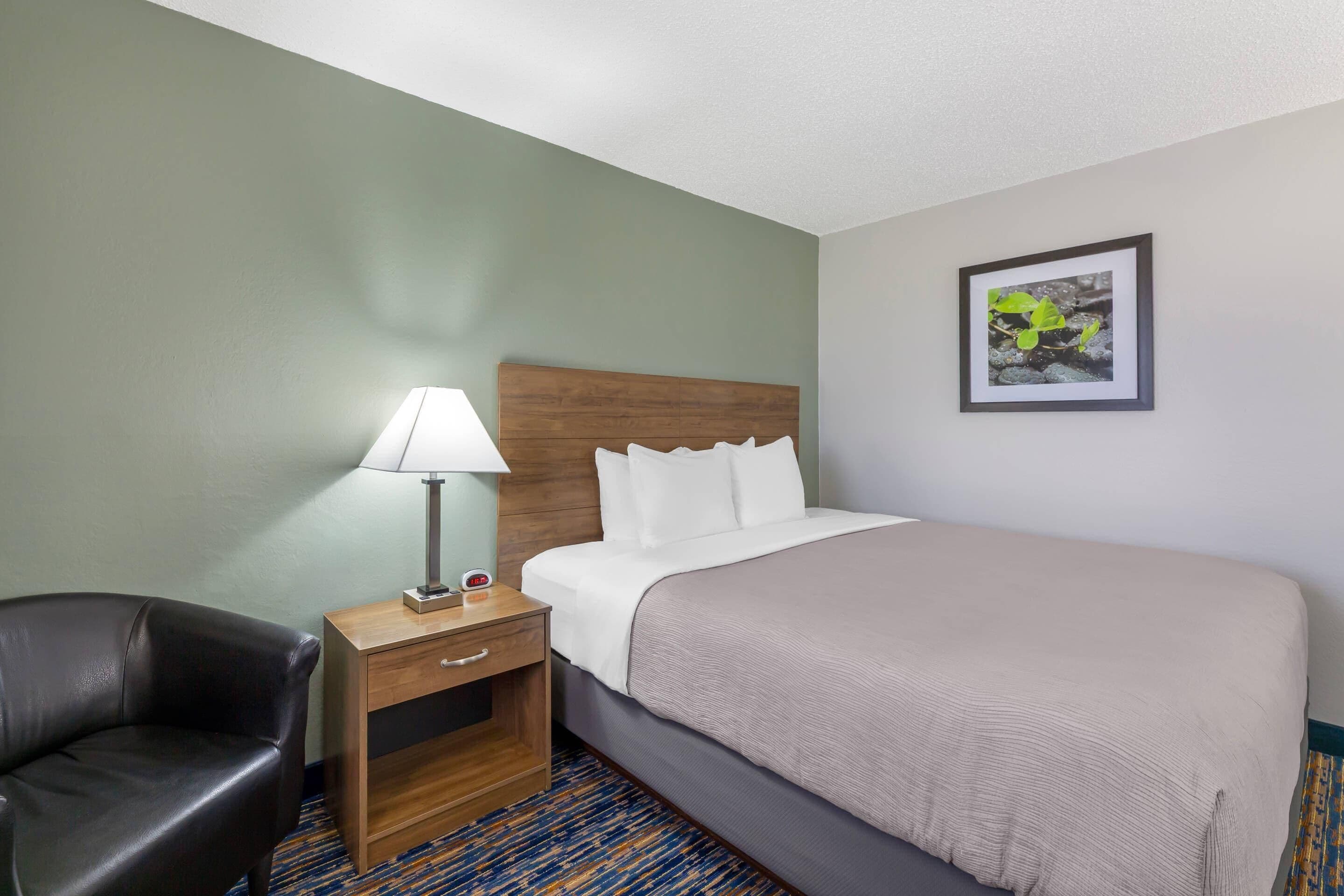 A hotel room with a bed, a nightstand with a lamp, a chair, and a framed picture on the wall.