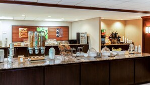 A hotel breakfast area with a self-service counter, coffee machines, and a variety of food items.