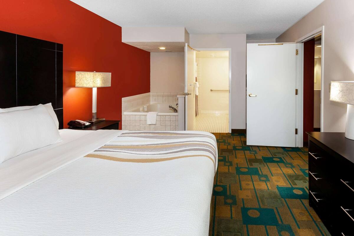 A hotel room with a bed, a nightstand, a lamp, a bathroom with a bathtub, and a patterned carpet.