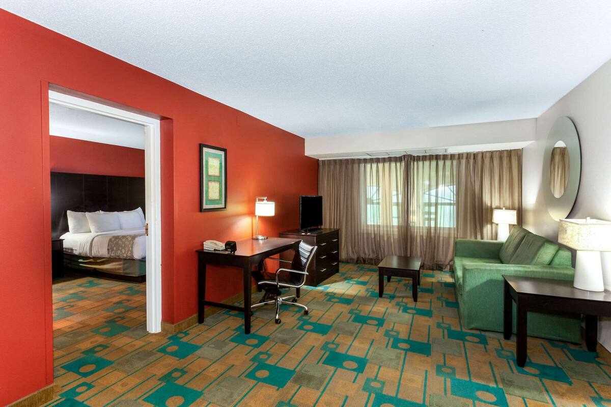 A hotel room with a red wall, a green sofa, a desk with a lamp, a television, and a bed with white bedding.