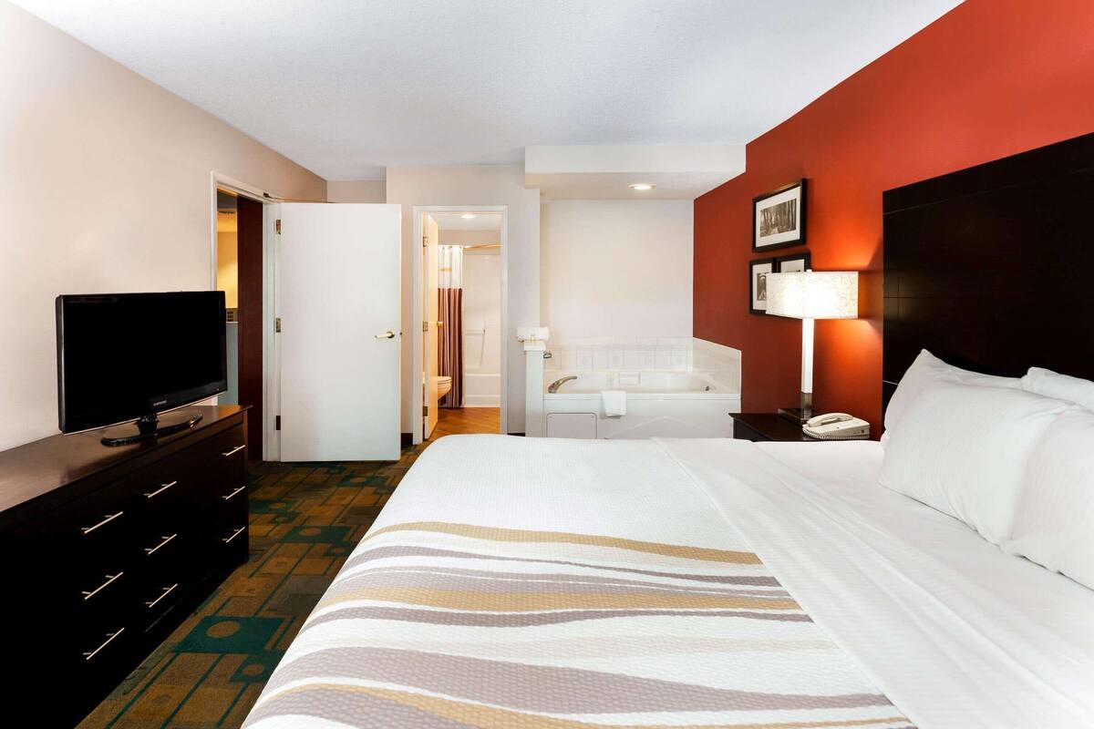 A hotel room with a bed, a television on a dresser, a bathroom with a bathtub, and a nightstand with a lamp.